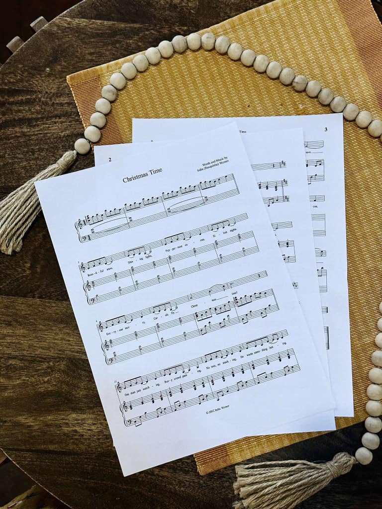 the Sheet music for "Christmas Time" by Lois and India laying on a table with a beige placemat and a string of beads.