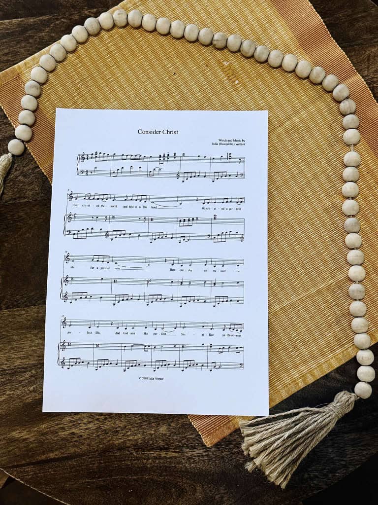 Sheet music to "Consider Christ" laying on a table with a beige placemat and a bead strand