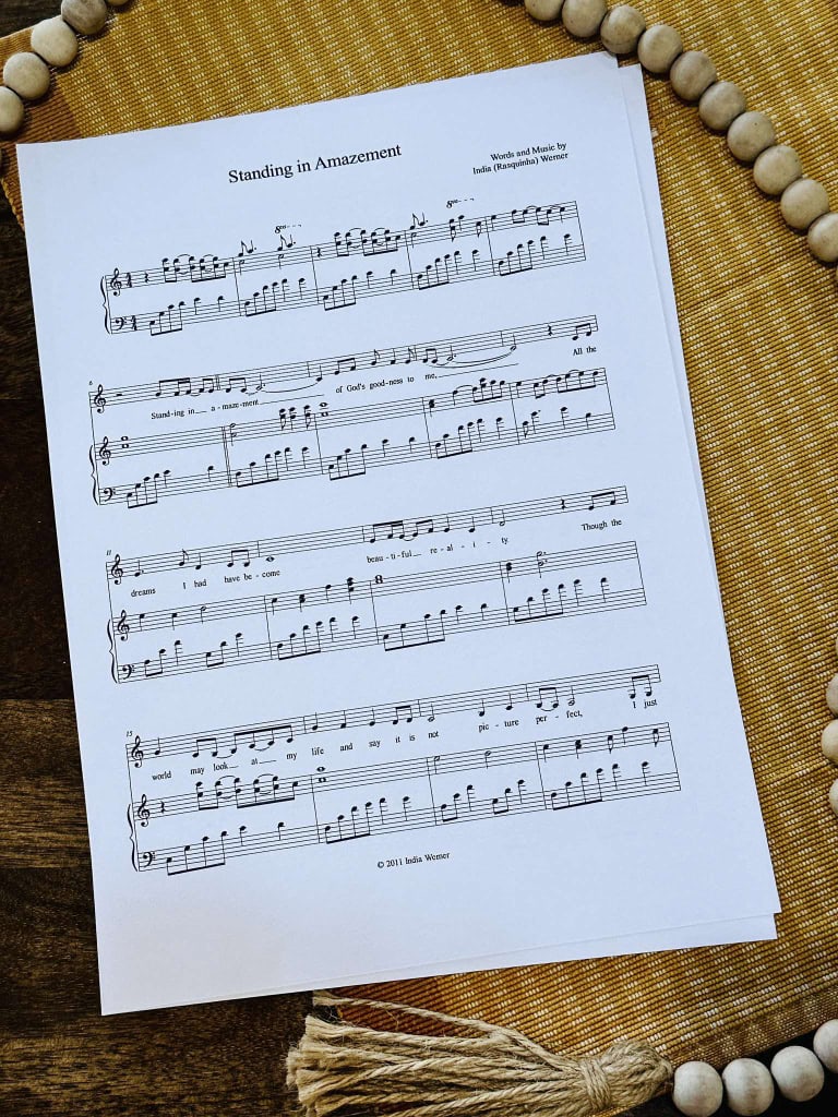 Sheet music to "Standing in Amazement" by Lois and India laying on a table with a beige placemat and a strand of beads.