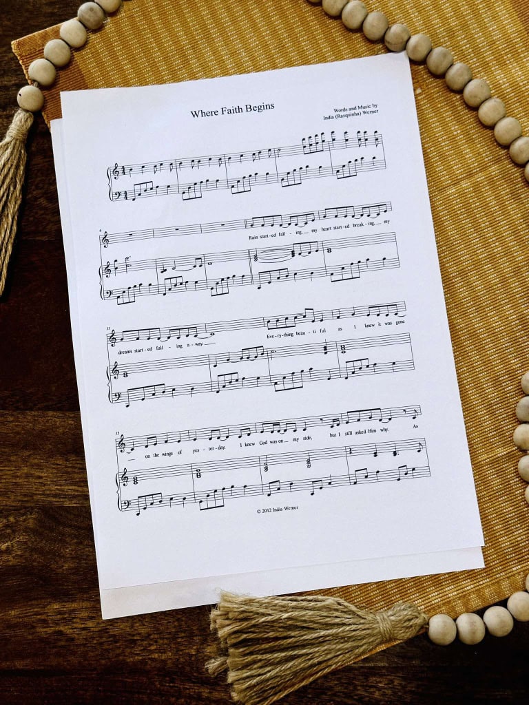 The sheet music to "Where Faith Begins" by Lois and India laying on a table with a beige placemat and a strand of beads.